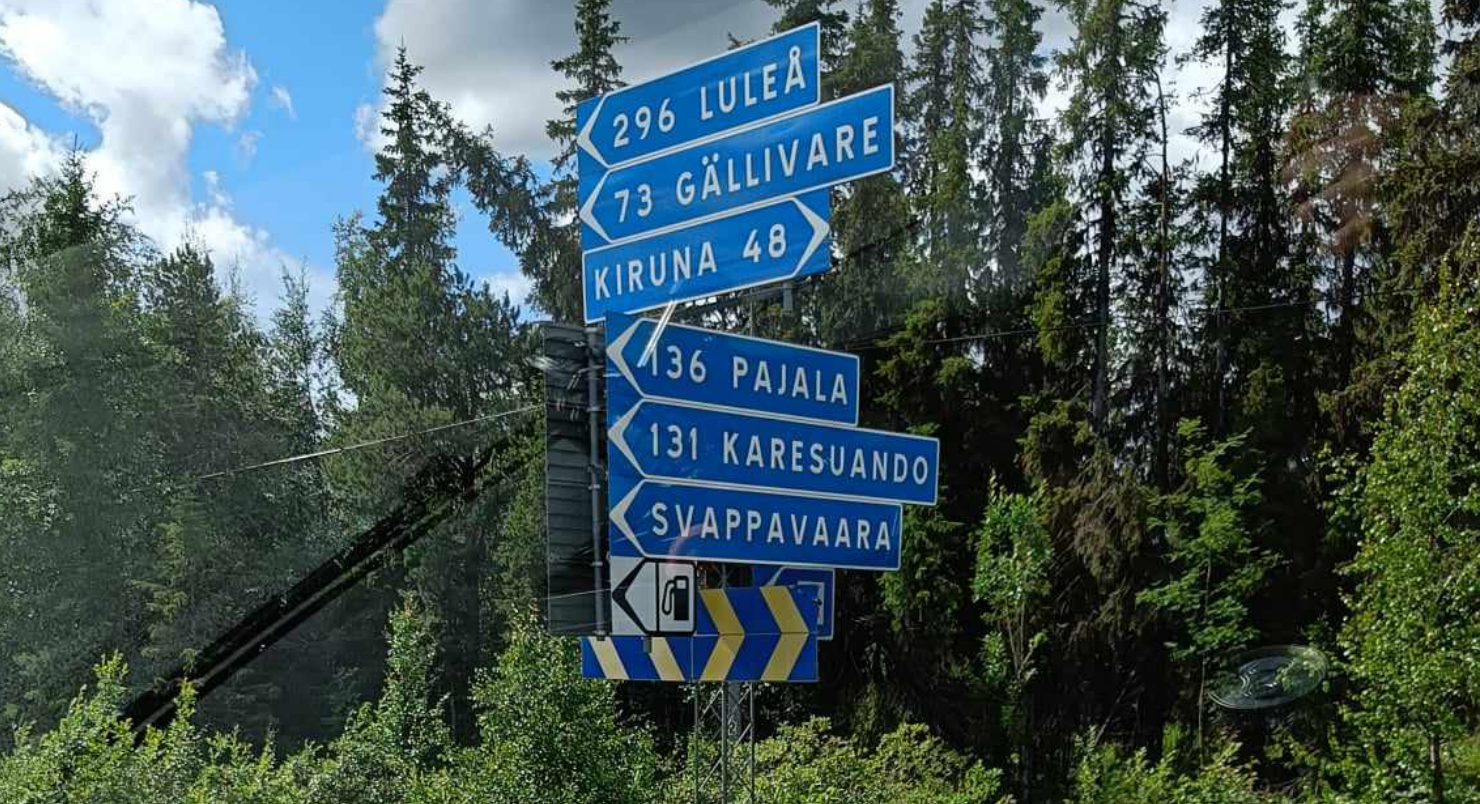 Skilt med stedsnavn og avstand foran en skog.